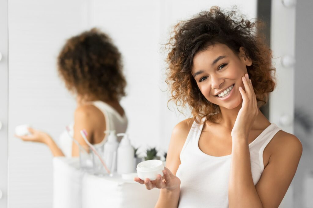 Afro Girl Using Face Cream Standing In Bathroom